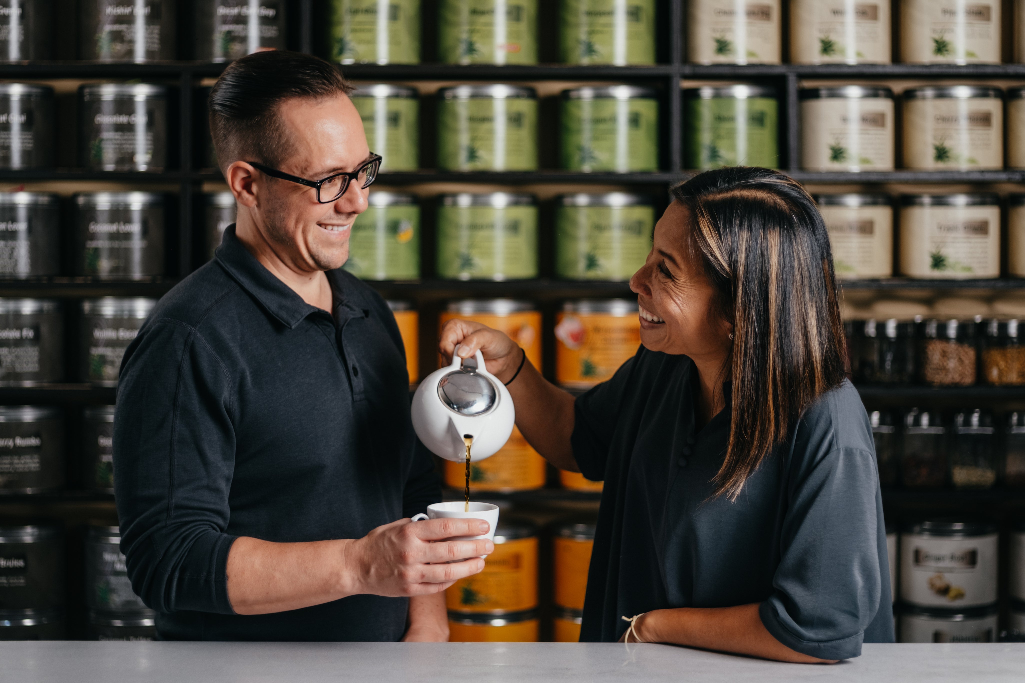 Two people in a tea shop with shelves of loose leaf tea canisters in the background